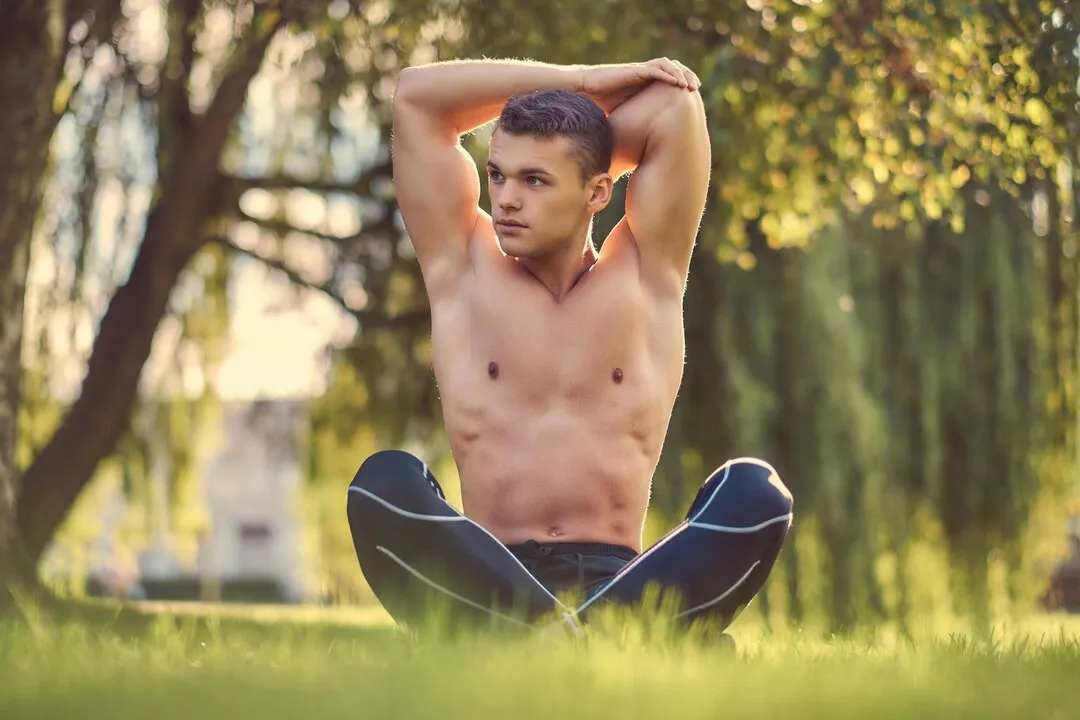 Shirtless young man practicing yoga in lotus pose on green grass, stretching hands and promoting a healthy lifestyle